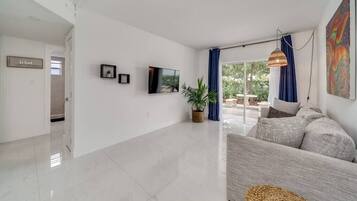 Un salon spacieux avec un grand canapé, un téléviseur à écran plat fixé au mur, une plante en pot et une vue sur l’extérieur grâce à de grandes fenêtres.