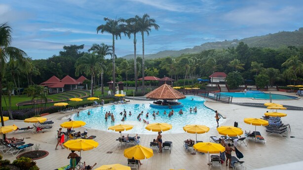 A large outdoor swimming pool surrounded by sun loungers and umbrellas, with palm trees and a hilly landscape in the background.