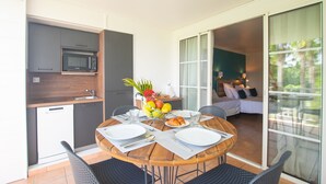 A compact kitchen with a microwave and a dishwasher, a dining table set for two with plates and cutlery, and a balcony with a bed and a view of greenery.