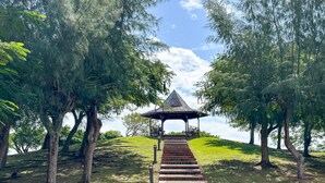 A gazebo with a sloped roof, surrounded by trees and a grassy hill.