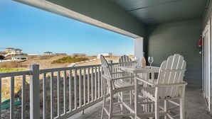 Un espace extérieur abrité, avec une table et des chaises, donnant sur une plage. On aperçoit une clôture et des bâtiments au loin.