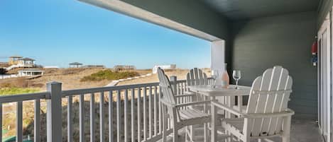 A covered outdoor seating area with a table and chairs, overlooking a beach with a fence and buildings in the distance.
