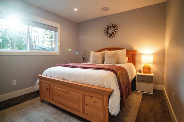 A bedroom with a wooden bed, a nightstand with a lamp, a window with blinds, and a wall-mounted wreath. - EV Charger, Hot tub, fire pit, family friendly (Stateline)