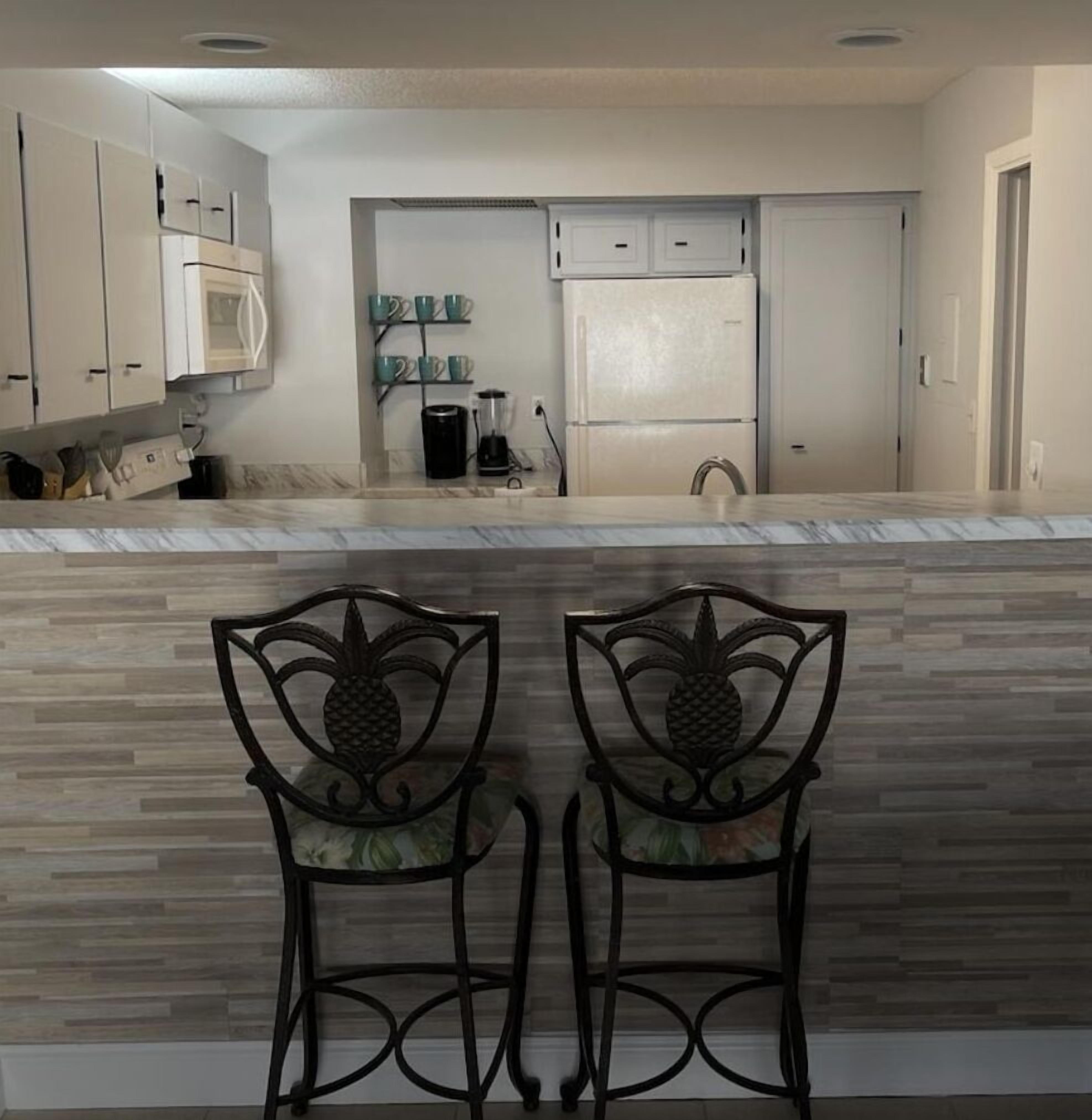 A kitchen with white cabinets, a double sink, and a countertop with a blender and coffee maker. There are two chairs with pineapple designs facing the kitchen.