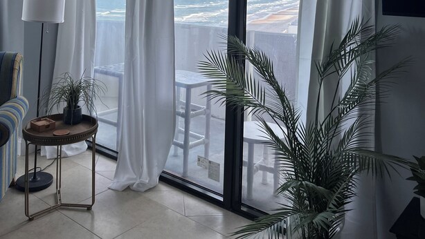 Ein Zimmer mit großem Fenster, weißen Vorhängen, einem kleinen Tisch mit Pflanze und einem Stuhl.