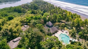 Ein tropisches Resort mit Schwimmbad, Hütten mit Reetdach und einem Strand mit Palmen.