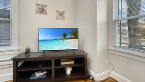Un salon avec un téléviseur à écran plat posé sur un meuble en bois foncé, deux petites étagères à plantes fixées au mur et une fenêtre avec des stores.
