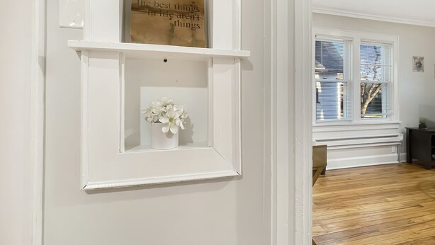 Une pièce avec un mur blanc, une étagère en bois ornée d’une citation et un vase blanc contenant des fleurs.