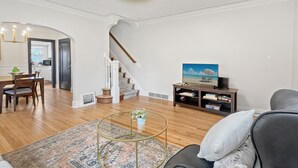 A spacious living room with a wooden floor, a round glass coffee table, a grey sofa, a television on a wooden stand, and a staircase leading to another room.