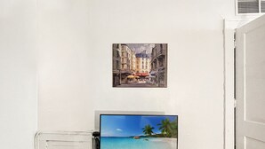 Habitación con televisor de pantalla plana sobre un mueble, un cuadro enmarcado que muestra una escena callejera y una puerta blanca.