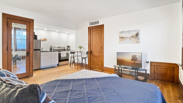 A bedroom with a large bed, a TV, and a kitchenette in the background.