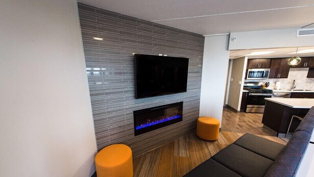 A modern living room with a flat-screen TV mounted on a tiled wall, a built-in fireplace, and a seating area with two orange ottomans and a black sofa. - Stay in the heart of downtown! Walk to Casey's Center, restaurants, and nightlife. Enjoy free parking and all the comforts of home. (Des Moines)