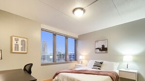 Une chambre d’hôtel comprenant un lit, un bureau, une chaise, deux tables de chevet, une lampe, un tableau encadré et une vue sur le paysage urbain grâce à de grandes fenêtres.