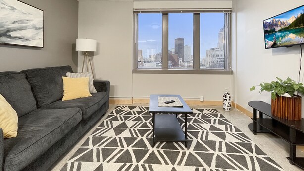 Un salon moderne avec un canapé gris, un tapis à motifs, une table basse et une grande fenêtre donnant sur la ville.