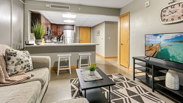 A modern living room with a sofa, coffee table, and TV stand. A kitchen is visible in the background with a refrigerator, microwave, and cabinets.