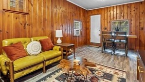 A room with wooden walls, a yellow sofa, a patterned rug, and a dining area with chairs and a table. - 1BR Residence With Vast Backyard (Des Moines)