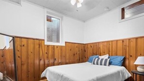 A bedroom with a wooden paneled wall, a bed with white bedding, a small bedside table with a lamp, and a window with blinds. - 1BR Residence With Vast Backyard (Des Moines)