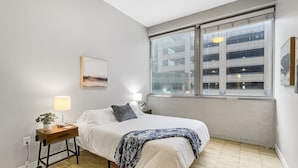A bedroom with a bed, bedside table, lamp, and a window with blinds. - Beautiful 2BR/2BA in Bldg connected to Skywalk (Des Moines)