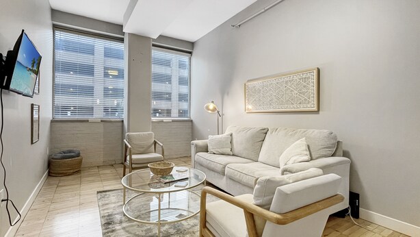 A modern living room with a sofa, armchair, and coffee table. There are two windows with blinds, a flat-screen TV mounted on the wall, and a framed artwork above the sofa. - Beautiful 2BR/2BA in Bldg connected to Skywalk (Des Moines)