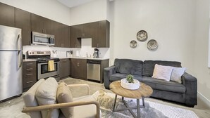 A modern kitchen with dark cabinets, a refrigerator, an oven, and a microwave. There is a gray sofa with cushions and a small wooden table with a decorative item.