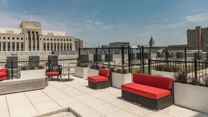 A rooftop terrace with outdoor seating, a view of a cityscape, and a large building in the background.