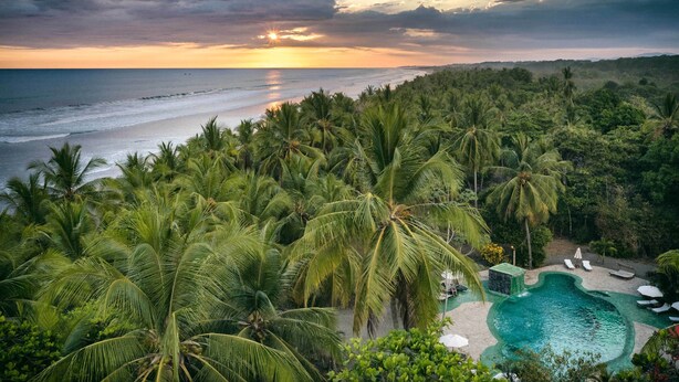 Un resort tropical con zona de piscina rodeada de palmeras y vista al océano al atardecer.