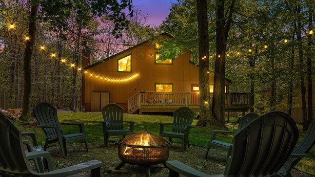 Ein gemütlicher Außenbereich mit Adirondack-Stühlen um eine Feuerstelle, einer Holzterrasse und Lichterketten.