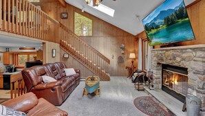 Ein geräumiges Wohnzimmer mit Kamin, Ledersofa, Holztreppe und einem großen Fernsehbildschirm.