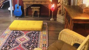 Een woonkamer met een gitaar, een bank, een vloerkleed met patroon, een rieten stoel en een beeld.