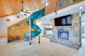 Un vaste espace de vie doté d’un plafond en bois, d’un toboggan vert en spirale, d’une cheminée en pierre et d’une cuisine située au fond de la pièce.