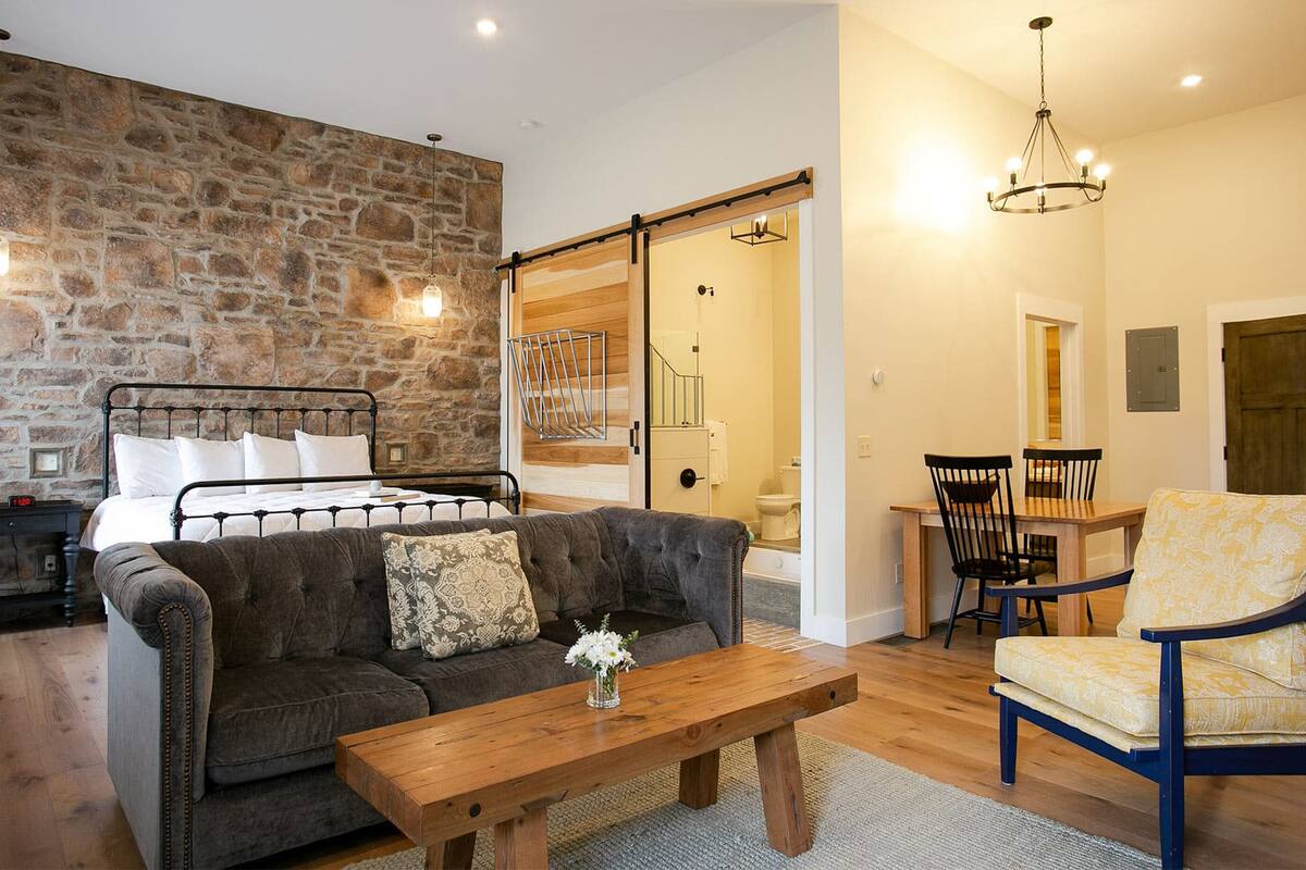 A cozy room with a stone wall, a bed, a sofa, a wooden coffee table, and a dining area with chairs and a chandelier.