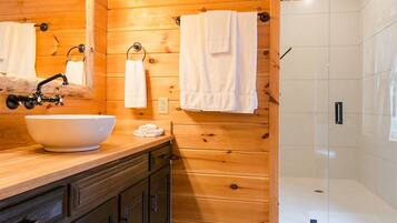Baño de cabaña de madera con un lavamanos grande, armarios oscuros, cerramiento de ducha de vidrio y paredes de madera.