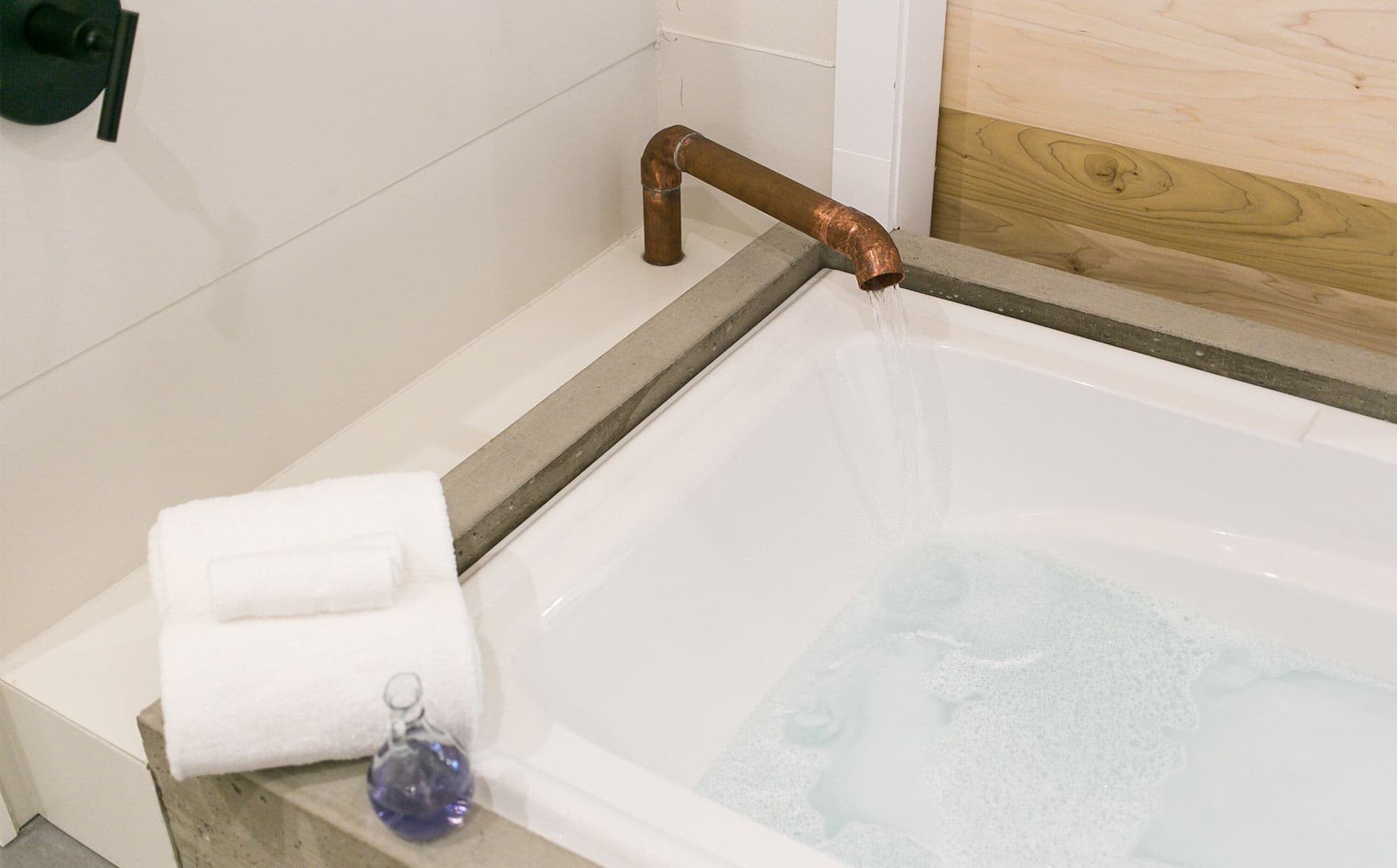 A bathtub with a copper pipe, a white towel, and a glass bottle on the edge.
