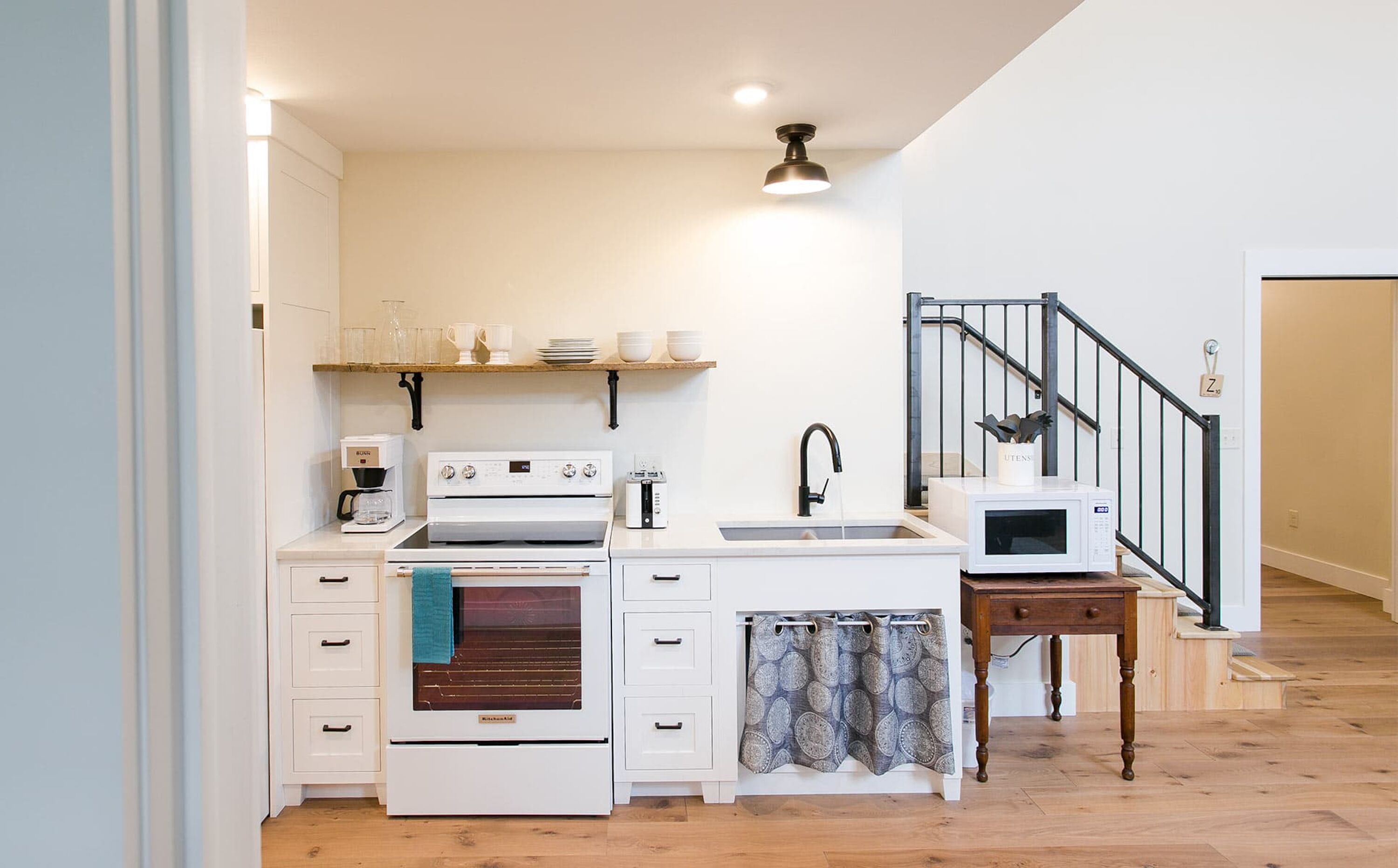 A compact kitchen with white cabinetry, a built-in oven, a microwave, and a coffee maker. There's a wooden shelf above the sink holding dishes and glasses. A patterned curtain hangs over the door, and a wooden table is next to the sink.