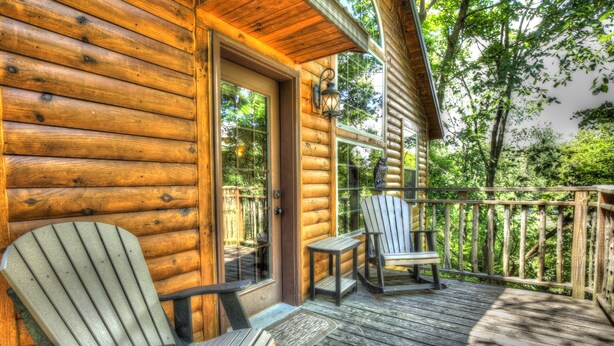 A wooden cabin with a porch featuring two Adirondack chairs and a small table. - Pine Cove Treehouses & Cabins (Millersburg)