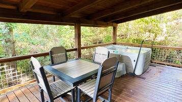 A covered outdoor area with a wooden table and chairs, a hot tub, and a view of the surrounding greenery.