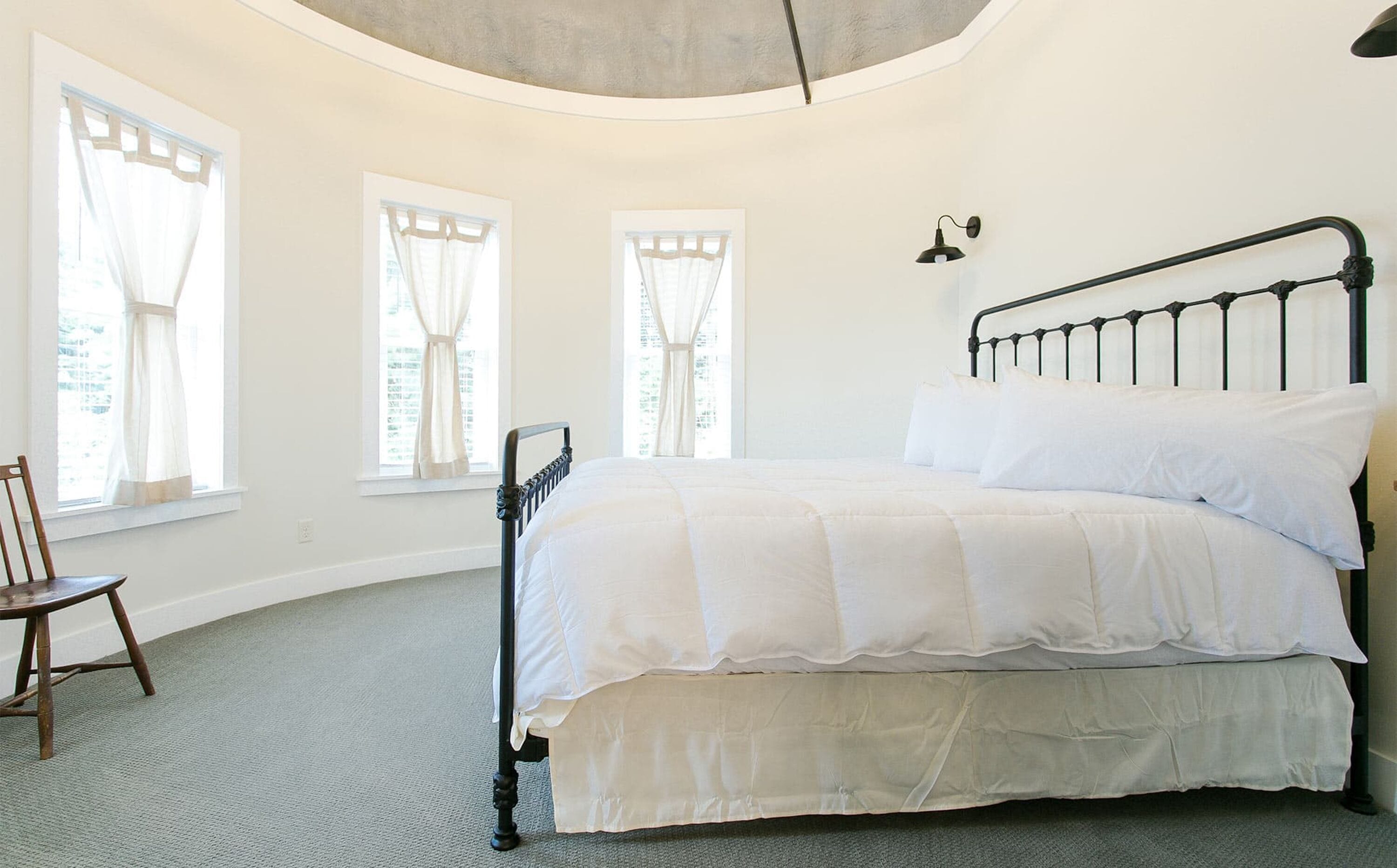 A bedroom with a bed, three windows, a chair, and a wall-mounted lamp.