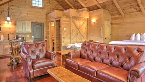 A log cabin interior with a wooden ceiling, a dining area with a wooden table and chairs, a leather sofa, and a bed with white bedding. - Pine Cove Treehouses & Cabins (Millersburg)