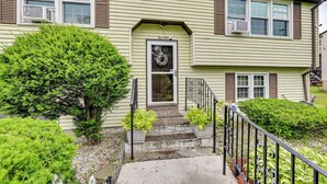 A two-story house with a front porch, a front door with a wreath, and a small garden area with potted plants. - Vintage Windsor Home: Private Yard & Covered Deck (Windsor)