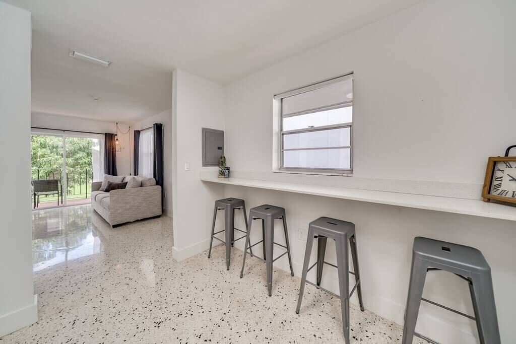 Un intérieur moderne avec un canapé blanc, trois tabourets de bar, une fenêtre avec des stores et une horloge au mur.