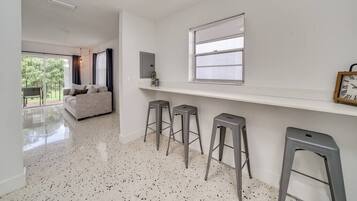 Un intérieur moderne avec un canapé blanc, trois tabourets de bar, une fenêtre avec des stores et une horloge au mur.