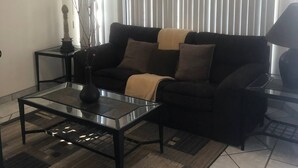 Un salon avec un canapé noir, une table basse en verre, un vase décoratif, une table d’appoint et une grande fenêtre avec des stores.
