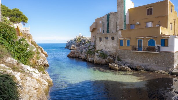 A coastal building with a stone facade, a balcony with blue shutters, and a clear view of the sea.