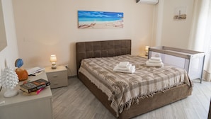 A bedroom with a large bed, a nightstand with a lamp, a painting of a beach, and a window with curtains.