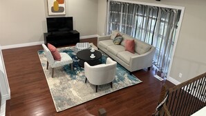 A living room with a sofa, two armchairs, a coffee table, a TV, and a large window with curtains.