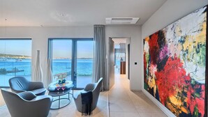A modern hotel room with a large window offering a view of the sea, a large abstract painting on the wall, and a seating area with a small table and two chairs.
