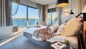 A modern bedroom with a large bed, a bathtub, and a view of the sea.