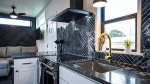 A modern kitchen with a black herringbone backsplash, a stainless steel range hood, and a gold faucet. - Contained In Gruene *Midnight* Spa/Outdoor Shower (New Braunfels)