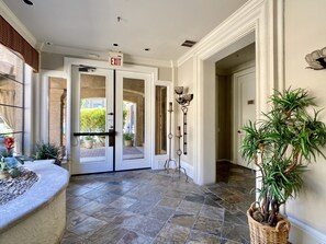 A hotel entrance with a stone-tiled floor, a glass door with an exit sign, a potted plant, and a wall-mounted light fixture.