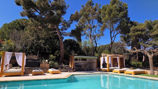A pool area with wooden loungers and a canopy bed, surrounded by trees and a building with a tiled roof.